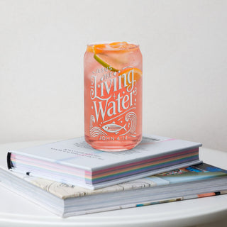 The Living Water can-shaped glass, pink iced drink on a stack of books, lettering facing forward.