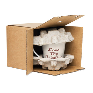 Cream-colored Love Thy Neighbor latte mug with burgundy script, inside protective cardboard packaging.