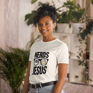 A woman wearing a white graphic t-shirt with the text 'NERDS FOR JESUS' and a book glasses design, standing in a brightly lit indoor room with plants.