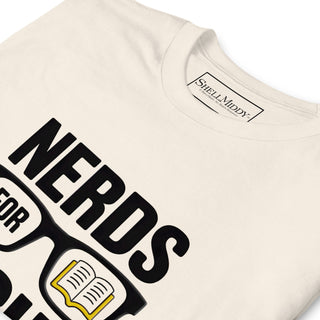 A close-up of a cream-colored t-shirt featuring a graphic print that reads 'NERDS FOR SHIT' in bold black letters, accompanied by a black eyeglasses graphic with an open book icon inside. The inside collar shows the brand label 'ShellMiddy'.
