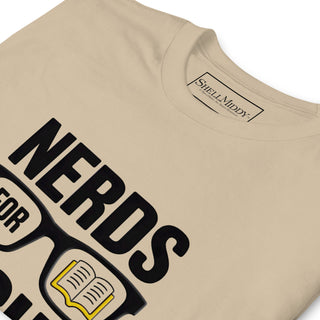 A close-up of a beige t-shirt featuring a graphic print that reads 'NERDS FOR SCIENCE' with glasses and an open book illustration. The inner collar label shows the brand 'Shell Middy'.