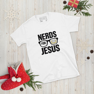 A white short-sleeve graphic T-shirt laid flat on a light wood surface with Christmas-themed props. The shirt has black text reading “NERDS FOR JESUS” with glasses and an open book graphic.