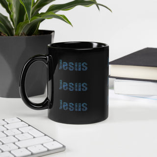 Black mug with 'Jesus' printed multiple times on a desk with a plant and books in the background