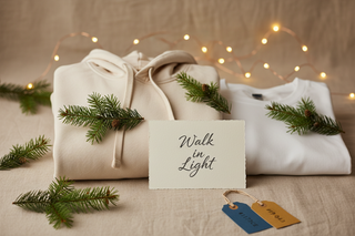 Festive but minimal holiday apparel scene featuring a cozy folded hoodie and t-shirt, small evergreen sprigs, a warm neutral background, and subtle gold holiday lights. Use ShellMiddy brand colors (#1173CE blue and #F8A434 gold) in small accents such as gift tags. No busy patterns; clean, premium composition. Include a tasteful Scripture card reading ‘Walk in Light’. Soft shadowing, warm glow, modern lifestyle photography, perfect for a Shopify signup popup.