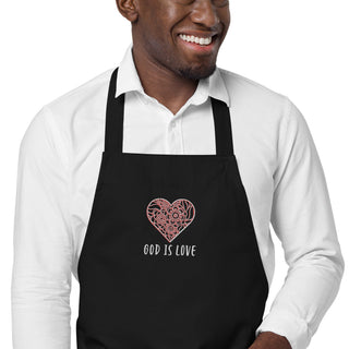 Close-up of floral heart embroidery and God Is Love lettering on a black apron worn by a model.