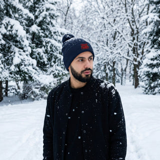 Model wearing a navy pom-pom knit cap with red and green Merry Christmas embroidery.
