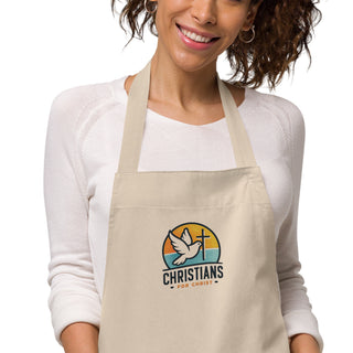 Close-up of dove-and-cross embroidery and Christians For Christ lettering on the beige apron worn by a woman.