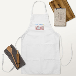 Hope + Works = Faith embroidered apron; white apron laid flat with cooking utensils.