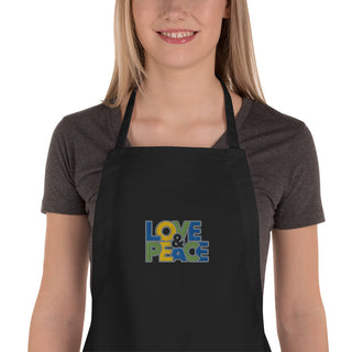 Love & Peace embroidered apron with blue, green, and yellow lettering, black apron on a model, chest close-up.