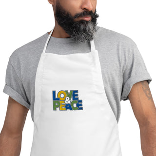 Love & Peace embroidered apron with blue, green, and yellow lettering, white apron on a model, chest close-up.