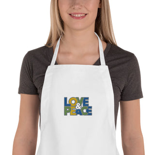 Love & Peace embroidered apron with blue, green, and yellow lettering, white apron on a model, chest close-up.
