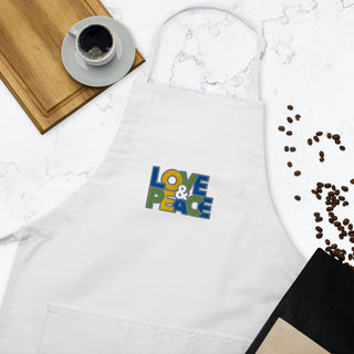 Love & Peace embroidered apron with blue, green, and yellow lettering, white apron beside coffee and kitchen props.