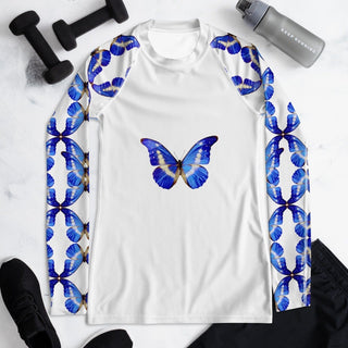 White long-sleeve rash guard with blue butterflies on the chest and sleeves, front laid flat beside exercise equipment